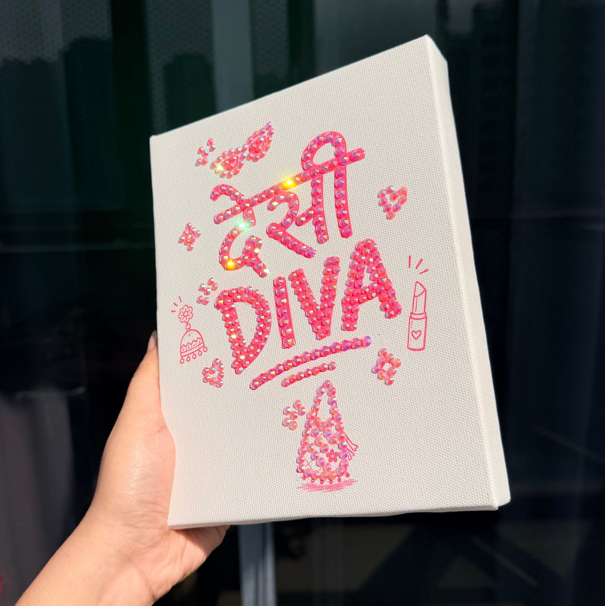 A hand holds a white rectangular canvas featuring pink Bedazzle Kit reading 'Desi Diva' with small rhinestones against a dark windowed background.